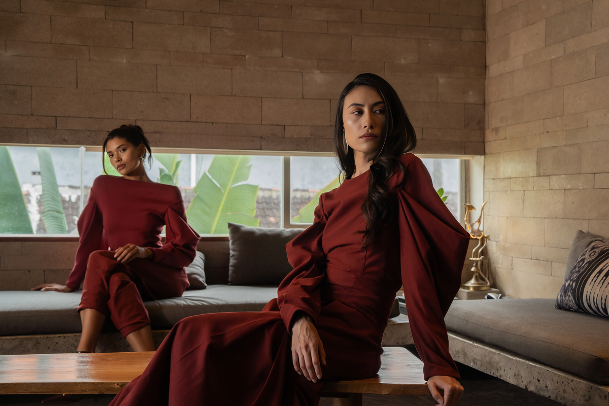 Two models in burgundy sculptural tailoring seated in a stone-walled room, Black Dahlia campaign