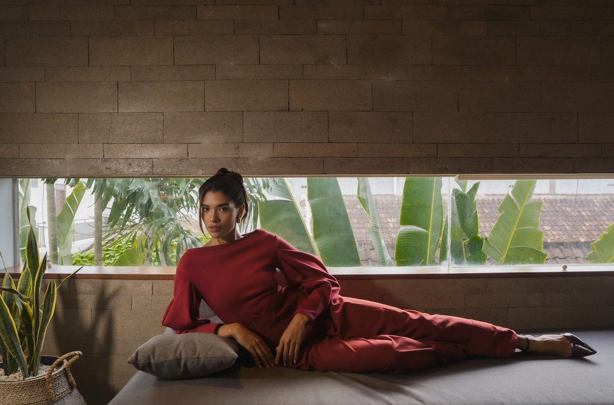 A model in burgundy tailoring reclining on a daybed beneath a window of banana leaves, Black Dahlia campaign