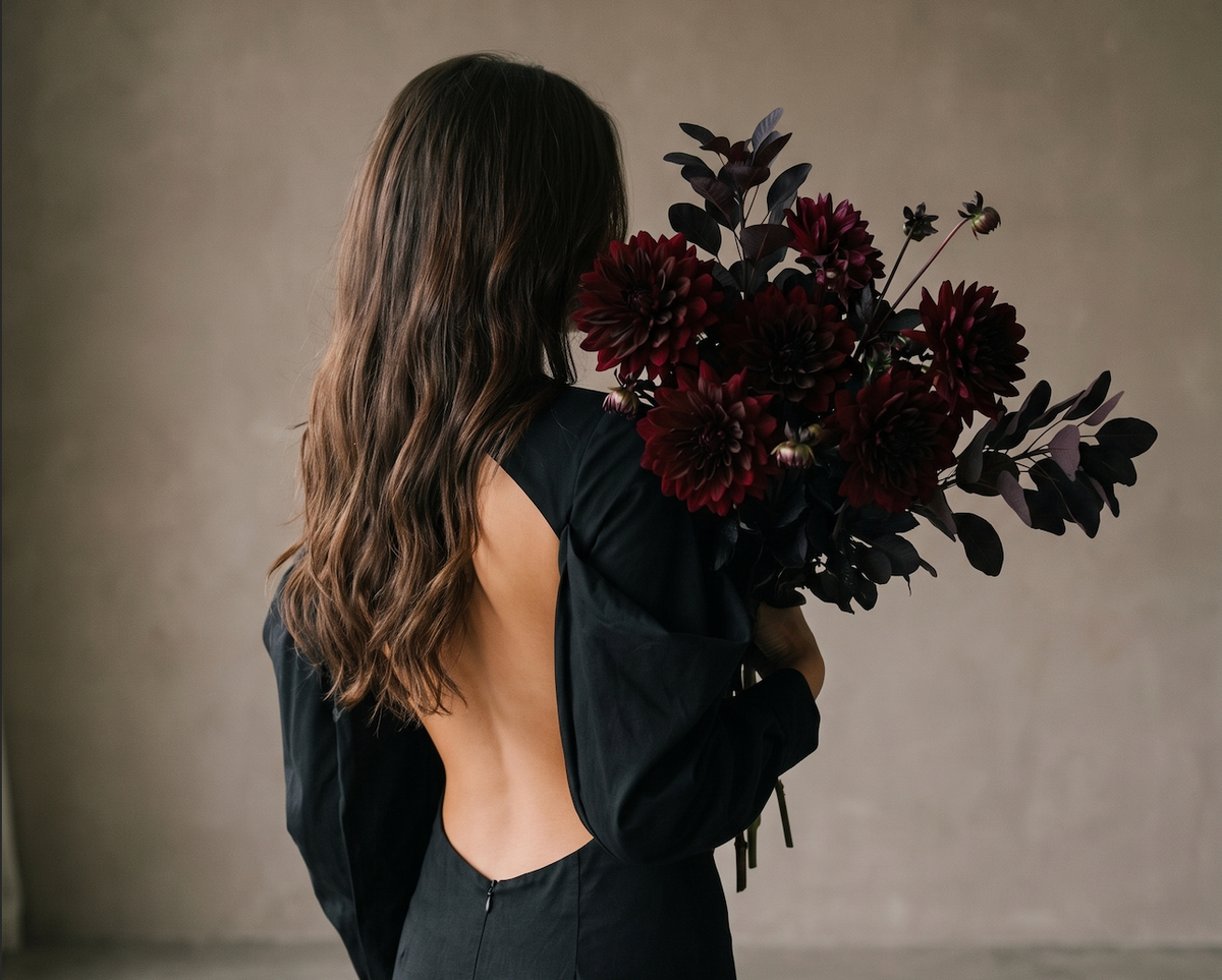 Model photographed from behind in a black open-back garment, holding a bouquet of dark burgundy dahlias, Black Dahlia Autumn/Winter 2026