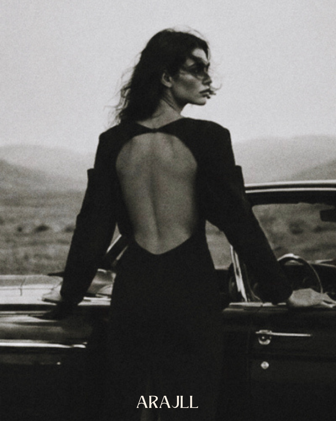 Black and white photograph of a model in a black open-back dress standing beside a vintage car in the desert, Black Dahlia campaign