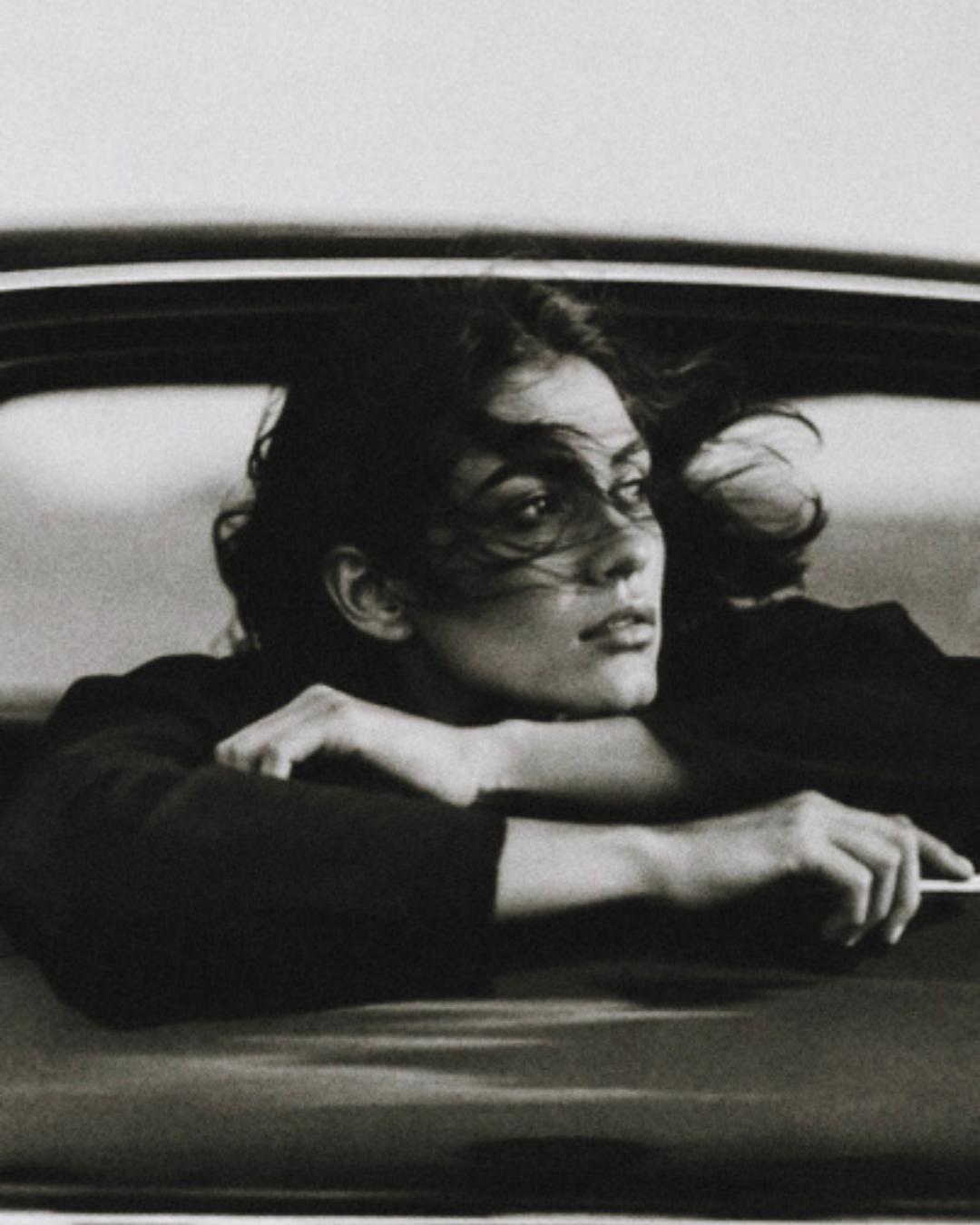 Black and white close-up of a model with wind-swept hair leaning on her folded arms through a car window, Black Dahlia campaign