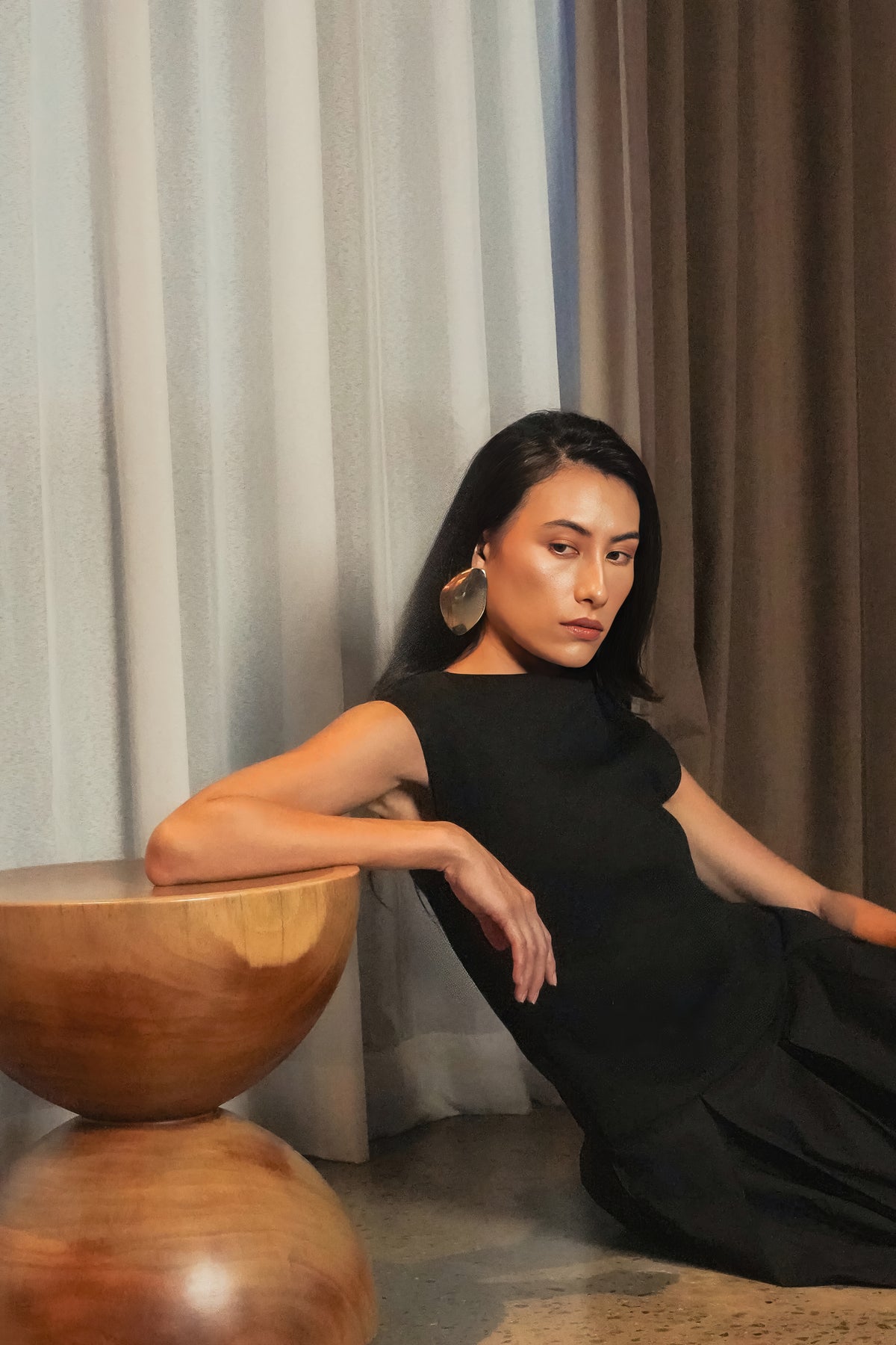 Model in a black sleeveless dress leaning against a round wooden side table, Black Dahlia campaign