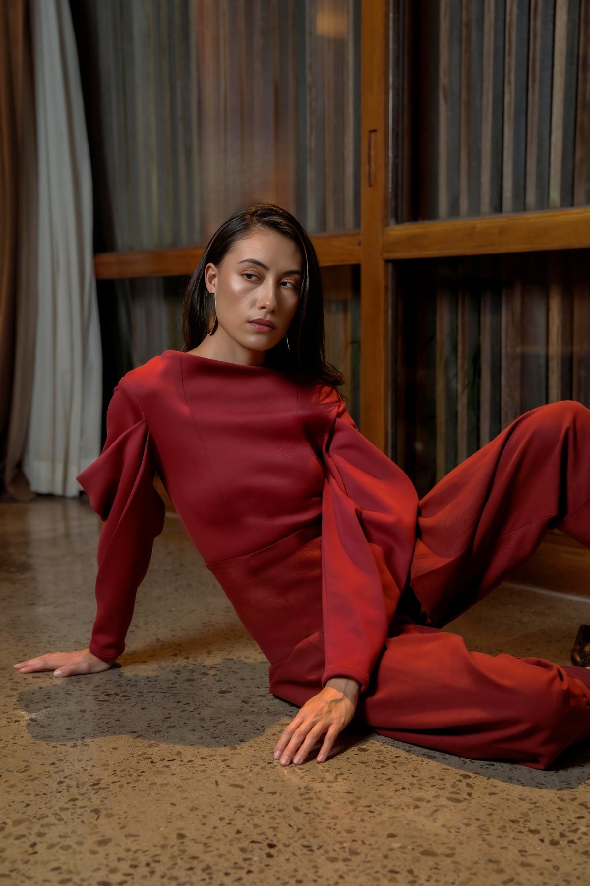 Model in a burgundy sculptural top and trousers seated on the floor with one leg raised, Black Dahlia campaign
