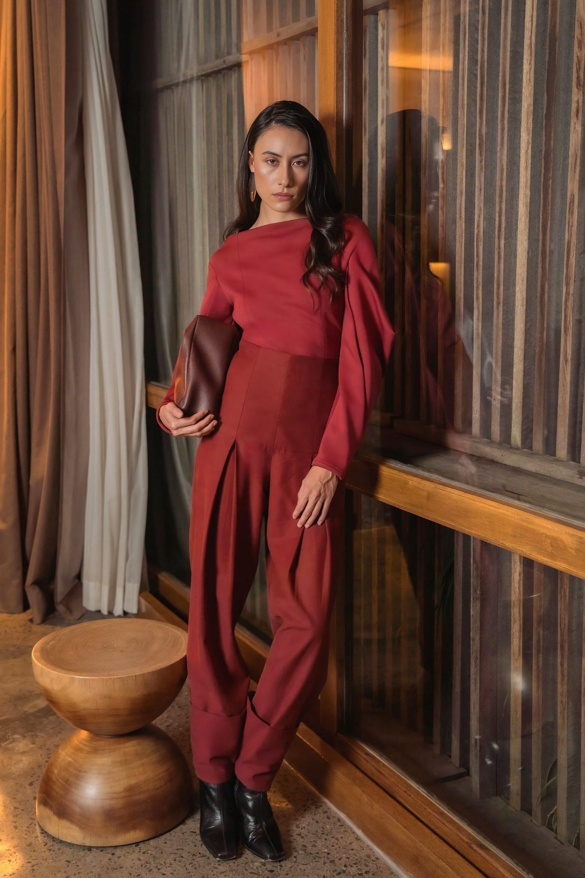 Model in a burgundy sculptural top and trousers beside a wooden-framed window, holding a brown clutch, Black Dahlia campaign
