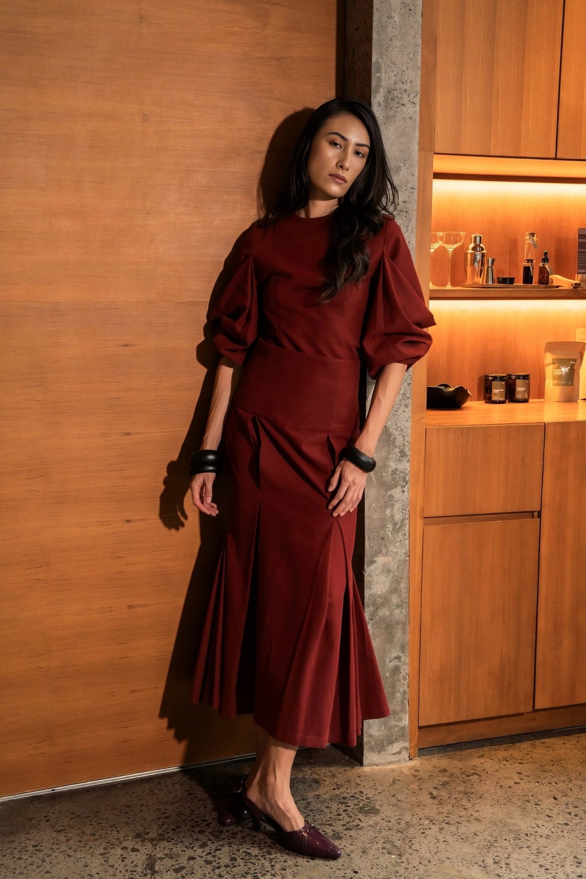 Model in a burgundy top with sculptural sleeves and a pleated skirt against a warm wooden wall, Black Dahlia campaign